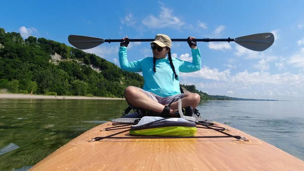 Tourist woman is paddling on a SUP board sunny summer day. Active sport recreation, outdoor exercising and lifestyle. Stand up paddle board with special seating chair, wet suit and life jacket.