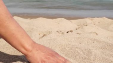 Young traveler on a sunny beach in exotic island gently stroking hot sand between her fingertips. Playful woman hand playing with yellow marine sand. Travel, adventure and leisure.