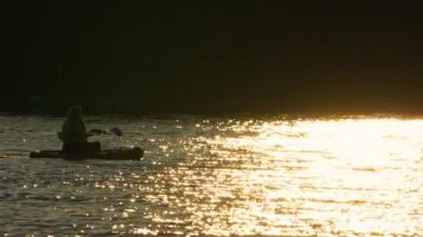 Bir kadının silueti SUP tahtasının üzerinde yelken açıyor. Gölün rahatlatıcı atmosferinin terapi amaçlı faydaları kavramı. Geri çekil ve iç sakinlik, ağır çekim. Geleneksel Kuzey Amerika su sporu.