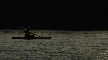 Bir kadının silueti SUP tahtasının üzerinde yelken açıyor. Gölün rahatlatıcı atmosferinin terapi amaçlı faydaları kavramı. Geri çekil ve iç sakinlik, ağır çekim. Geleneksel Kuzey Amerika su sporu.