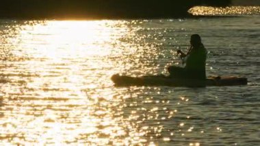 Bir kadının silueti SUP tahtasının üzerinde yüzüyor, altın saat günbatımında kürek çekiyor. Manitou Gölü 'nde hareketli eğlence ve spor için ayağa kalkın. Aktif tatil, meditasyon ve rahatlama.