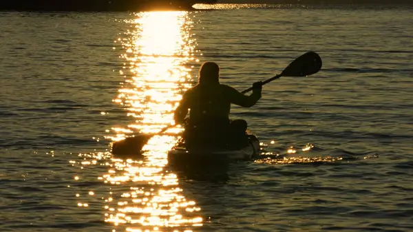 Bir kadının silueti SUP tahtasının üzerinde yüzüyor, altın saat günbatımında kürek çekiyor. Manitou Gölü 'nde hareketli eğlence ve spor için ayağa kalkın. Aktif tatil, meditasyon ve rahatlama.