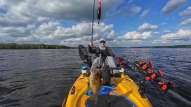 Güneş kremi kapüşonlu ve şapkalı balıkçı, kano kullanırken kameraya kendini gösteriyor. Güneşli bulutlu bir günde gölün ortasında tek başına vakit geçirmek. Kamerada kaya levreği ve balon çizgisi