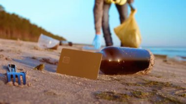 Plajdaki kredi kartı ve plastik atıkların birleşik sahnesi, plastik atık sorununu ve bunun azaltılması gereğini vurguluyor. Tüketici Sorumluluğu kavramı, Plastik Atık