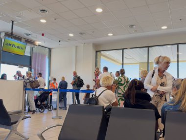 Gezginler, hava baltık uçuşları için hareketli bir terminalde toplanarak güneşli bir günde yolculuklarına hazırlanan çocuklu ve bireyli aileleri vurguluyorlar..