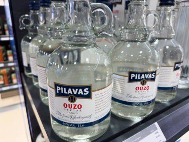 Birkaç şişe Pilavas uzo bir mağaza rafında sergileniyor, her birinde açık bir sıvı karışımı var. Perakende ortamı bu geleneksel Yunan ruhunu vurguluyor.