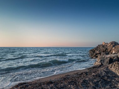 Dalgalar kumlu sahile yuvarlanıyor, güneş batarken kayalık oluşumlarla karşılaşıyor, gökyüzüne yumuşak renkler saçıyor ve huzurlu bir atmosfer yaratıyor..