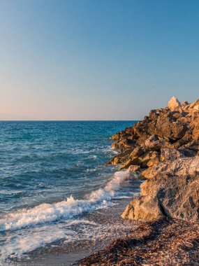 Gün batımında, dalgalar yavaşça kayalık bir kıyı şeridine çarpar, gökyüzünün ılık renklerini yansıtarak, deniz kenarında huzurlu bir atmosfer yaratır..