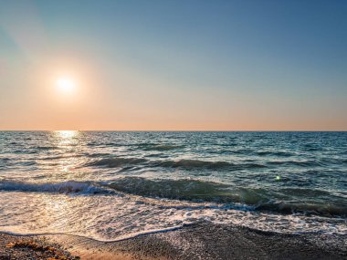 Güneş ufuktan batıyor, sakin okyanus sularına sıcak bir parıltı saçıyor. Yumuşak dalgalar kumlu sahile vuruyor ve akşam ışığında huzurlu bir atmosfer yaratıyor..