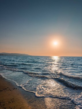 Batan güneşin ılık parıltısı sakin okyanusa yansıyor kumlu sahilde yumuşak gölgeler bırakıyor ve hafif dalgaları vurguluyor. Uzak tepeler bu huzurlu manzarayı tamamlıyor..