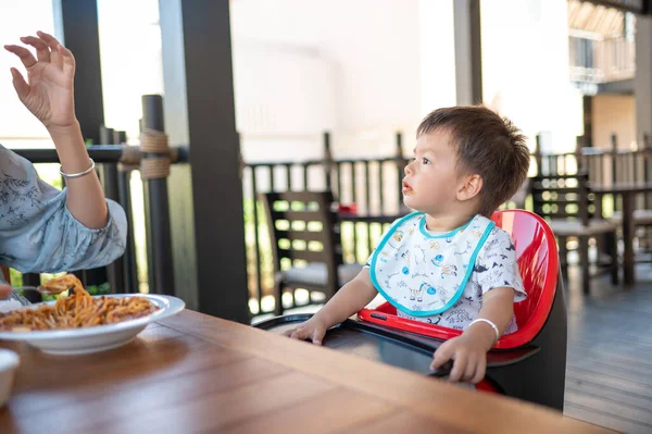 Toddler sitting on a high chair in the restaurant during a meal. Handsome multiracial one and half year old baby boy having a dinner meal in the restaurant with his mother feeding him in an unrecognizable restaurant at evening