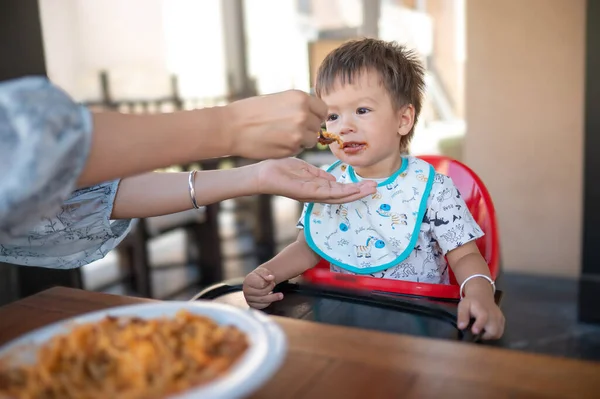 Toddler being fed in the restaurant by his mom. Handsome multiracial one and half year old baby boy having a dinner meal in the restaurant with his mother feeding him in an unrecognizable restaurant.