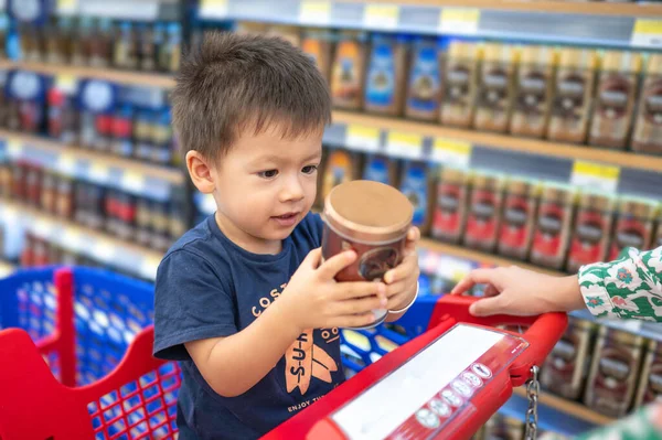 Two year old adorable multiracial toddler in a blue t-shirt sitting in a shopping trolley with groceries, holding a plastic cup with small hands, in grocery store