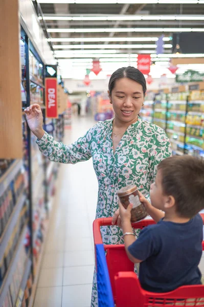 Young and smiling asian mom in green floral dress and her little multiracial boy in shopping trolley buying groceries in supermarket