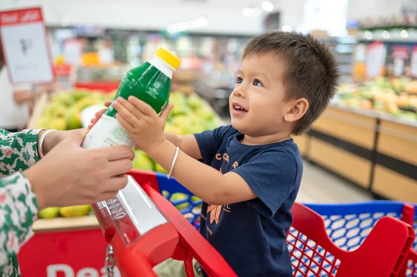 Two year old adorable, smiling multiracial toddler in a blue t-shirt sitting in a shopping trolley with groceries, holding a bottle with small hands, in grocery store