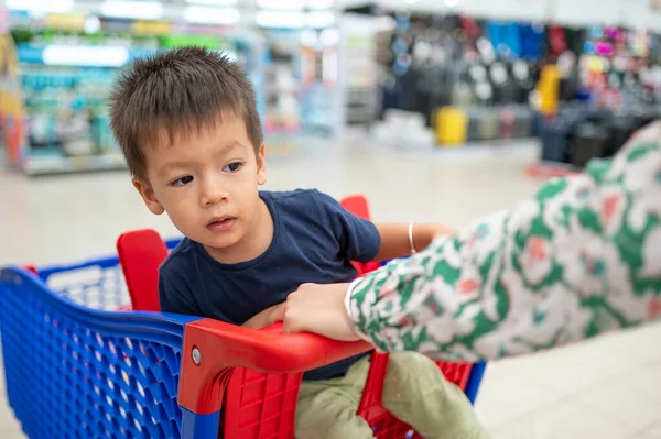 Two year old adorable multiracial toddler in a blue t-shirt sitting in a shopping trolley with groceries, in grocery store
