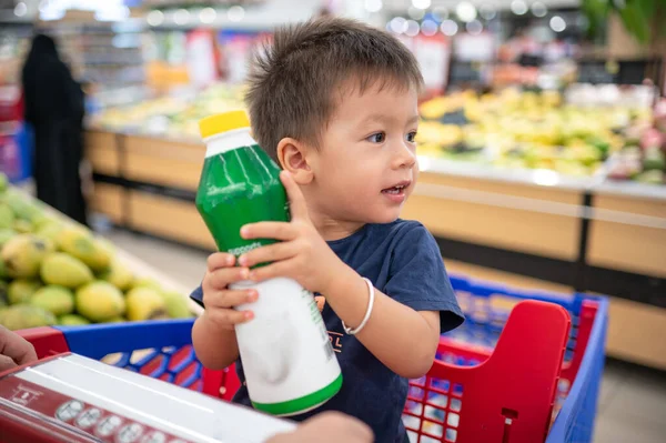 Two year old adorable, smiling multiracial toddler in a blue t-shirt sitting in a shopping trolley with groceries, holding a bottle with small hands, in grocery store