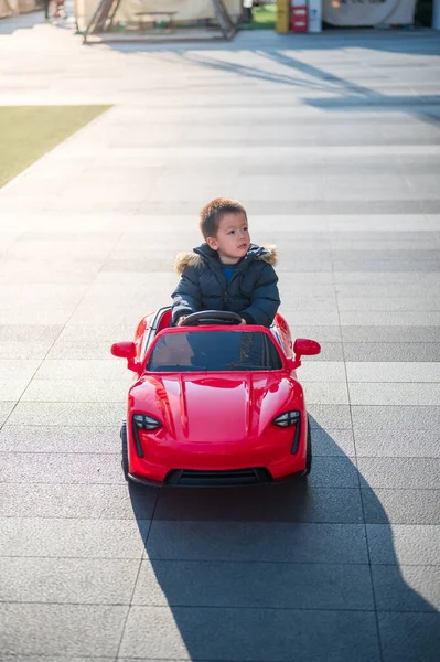 一位年轻的多种族幼儿开着一辆红色的运动玩具车快乐地在街道上穿梭 他们穿着冬季夹克 控制着自己的玩具车 脸上挂着大大的笑容 图库图片