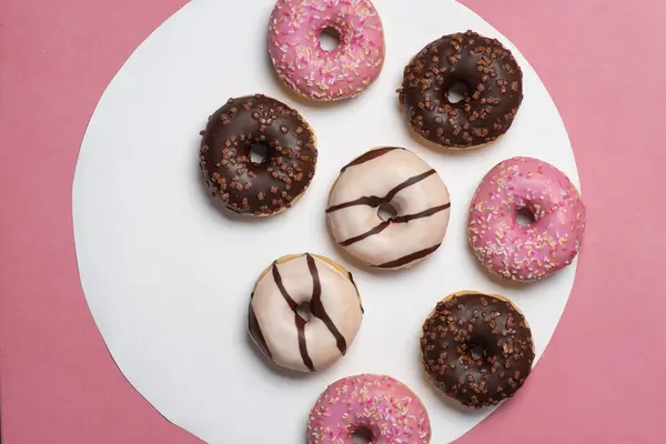 Stock image A delightful assortment of donuts with vibrant pink, chocolate, and white icing, paired with sprinkles, showcased on a contrasting white and pink backdrop. Perfect for pastry lovers.