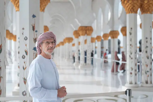 stock image A man dressed in traditional attire stands inside Sheikh Zayed Grand mosque in Abu Dhabi, surrounded by ornate columns beautifully decorated with gold and floral patterns