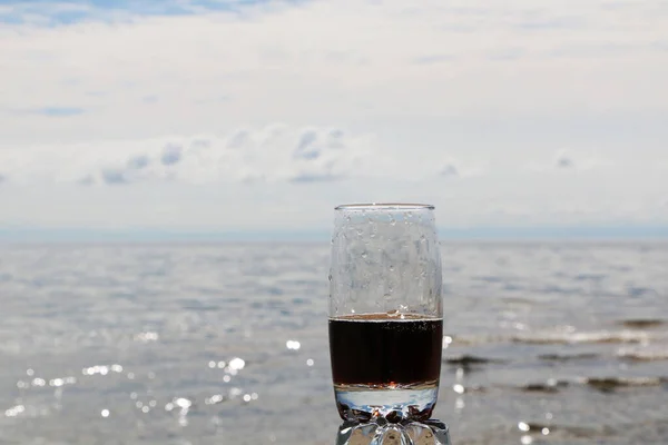 Brown carbonated drink in a beautiful glass at sunset by the sea and in a cafe. Cold frothy drink in the heat on the beach.