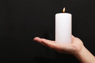 Bir adam elinde yanan beyaz bir mumu siyah bir arka planda tutuyor. Siyah bir arka planda elinde bir mum yanıyor..