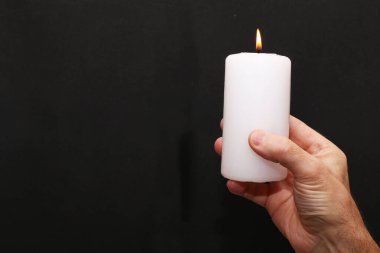 Bir adam elinde yanan beyaz bir mumu siyah bir arka planda tutuyor. Siyah bir arka planda elinde bir mum yanıyor..