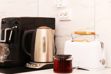 Bir adam ev aletlerini kullanarak kahvaltı hazırlar. Ekmek kızartma makinesi, çaydanlık ve kahve makinesi masanın üstünde..