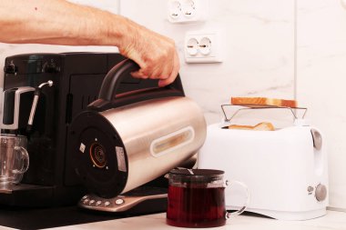 Bir adam ev aletlerini kullanarak kahvaltı hazırlar. Ekmek kızartma makinesi, çaydanlık ve kahve makinesi masanın üstünde..