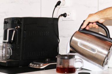 Bir adam ev aletlerini kullanarak kahvaltı hazırlar. Ekmek kızartma makinesi, çaydanlık ve kahve makinesi masanın üstünde..