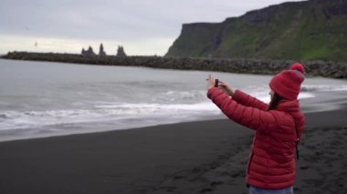 Kız telefonda okyanusun resimlerini çekiyor. Kız siyah bir plajda duruyor. İzlanda.