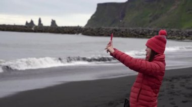 Kız telefonda okyanusun resimlerini çekiyor. Kız siyah bir plajda duruyor. İzlanda.