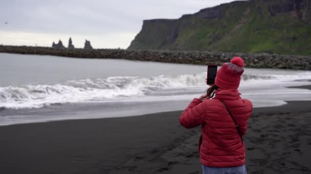 这个女孩在电话里给大海拍照 这个女孩站在一个黑色的海滩上 — 图库视频影像