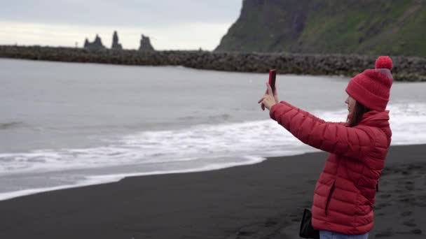 その少女は電話で海の写真を撮る 少女は黒いビーチに立っている アイスランド — ストック動画