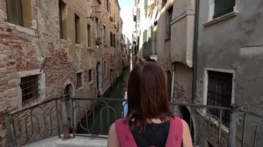 Venedik. Bir turist köprü boyunca geriye doğru yürür ve dar bir kanalın arka planına gülümseyerek döner..
