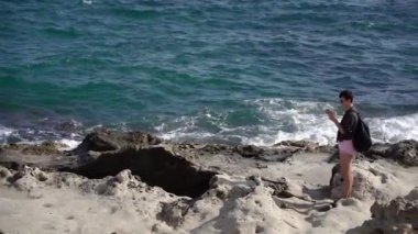 Bir kız denizin arka planında kayalık bir sahilde duruyor ve telefonuna bakıyor..