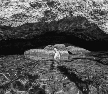 Bir kadın berrak suda bir kayanın altında duruyor ve gülümsüyor..