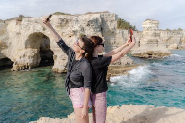 İki kız sırt sırta durarak denizin arka planına karşı dururlar ve telefonlarıyla kendilerini filme alırlar..