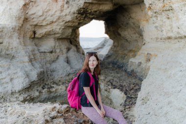 Bir kadın kayaya oturur ve kayalık bir sahile bakar..