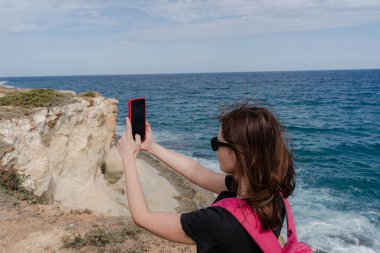 Bir kadın telefonuyla resimli bir plajın fotoğrafını çekiyor. Kayaların güzel manzarası.