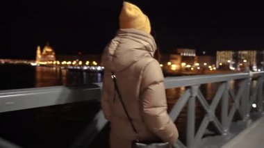 Bir kadın bir gece şehrinin arka planına karşı köprüden geçiyor..