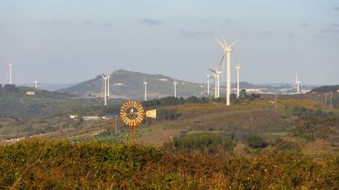 Bulutlu bir gökyüzünün altında, yel değirmenli yeşil bir tepedeki rüzgar türbinlerinin manzarası.