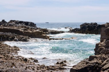 Sakin deniz manzarası, dalgalar kayalık kıyılara çarpıyor berrak mavi bir gökyüzü altında, görülebilir bir tekneyle