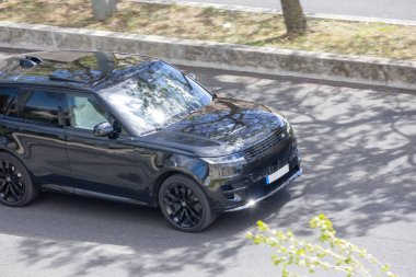 Siyah Range Rover güneşli bir günde sürüyor, pencerelerdeki ağaçları yansıtıyor.