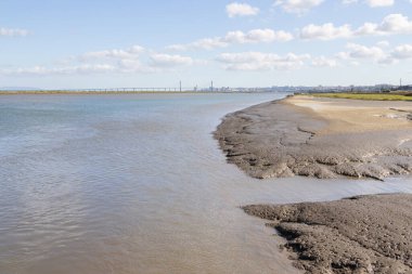 Tagus nehrinin alçak gelgitte Atlantik okyanusuna akan manzarası, bir köprü ve arka planda Lizbon şehri.
