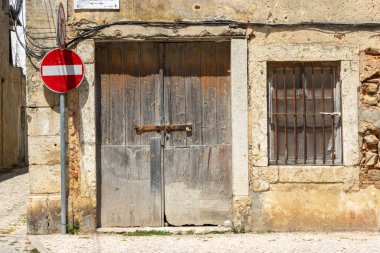 Paslı kilidi ve demir parmaklıklı penceresi olan eski, yıpranmış ahşap kapı, ön planda giriş işareti olmayan taş bir duvara yerleştirilir.