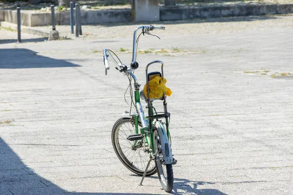 stock image Green folding bike with a yellow plastic bag on the rack is standing on a paved square on a sunny summer day