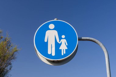 Yuvarlak mavi yaya işareti, arka planda açık mavi gökyüzü olan bir yetişkinin ve bir çocuğun el ele tutuşmasını gösteriyor.