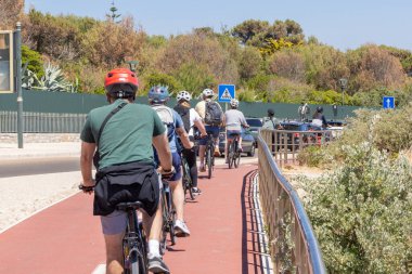 Bir grup arkadaş, güneşli bir yaz gününde bisiklet yolunda kask takarak bisiklet sürüyor.