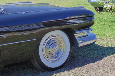 Klasik siyah Cadillac Deville. Çimlerin üzerine park etmiş. Ön tekeri beyaz lastik ve krom detaylarıyla gösteriyor.